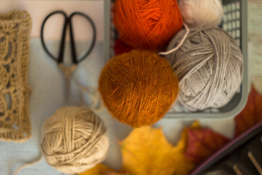 Cozy autumn flatlay with orange and gray wool yarn balls, vintage scissors, crochet lace, and fall leaves. Warm flat lay photograph for needlework and craft hobby concepts.