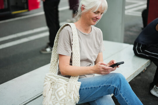 Caucasian senior woman using smartphone at bus station wearing casual clothes - City lifestyle and technology concept - Main focus on face - UGC content
