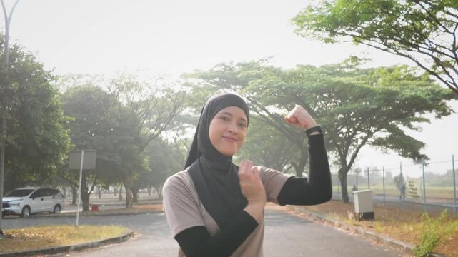 A confident Asian Muslim woman flexes her arm during an outdoor fitness session in a sunlit park, expressing strength, empowerment, healthy lifestyle and determination.