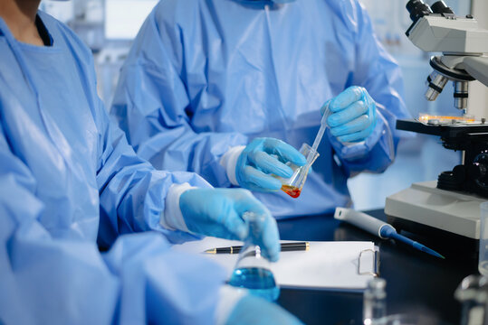 Two dedicated medical scientists in surgical protective gowns using a micropipette and microscope in research laboratory.