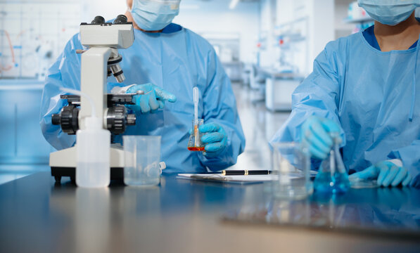 Close up of medical researchers in blue protective gowns using a pipette and beaker scientific testing near a microscope in a laboratory.