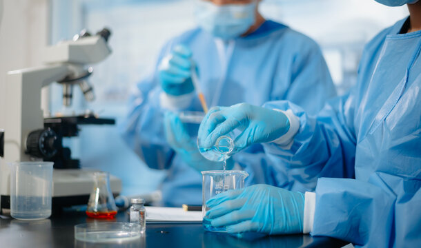 Close up of medical researchers in blue protective gowns using a pipette and beaker scientific testing near a microscope in a laboratory.