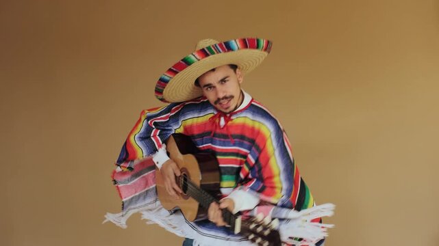 Young adult man wearing straw sombrero and vibrant striped serape holds acoustic guitar upright while facing camera, both hands on instrument and torso turned toward viewer indicating active