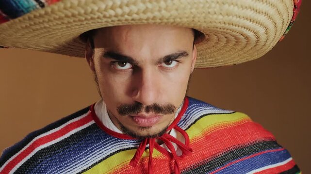 Frontal studio portrait shows adult man in large woven straw hat and striped serape lowering brim while tilting head down, revealing deeper brow furrows, rugged features, intense gaze, moody