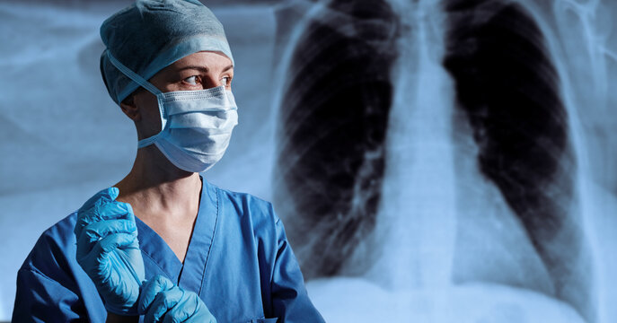 Female surgeon in protective mask and scrub cap adjusting sterile gloves in front of a chest radiography display before starting thoracic surgery.