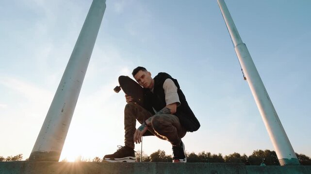 Low-angle outdoor scene shows young man crouching with skateboard on concrete ledge, framed by two tall poles against clear sky, with skateboard, skater, young, man detail, showing crouching, outdoor