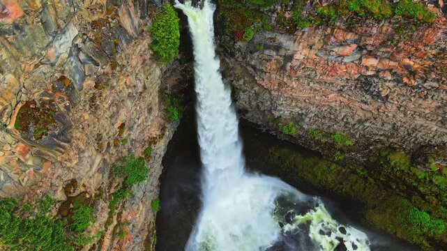 Aerial high angle shot of a powerful waterfall plunging down a steep rocky canyon cliff into a pool surrounded by lush green foliage on daylight