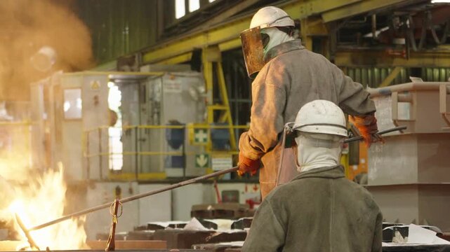 Steel foundry workers in heavy safety gear tend molten metal with long tools inside an industrial workshop.