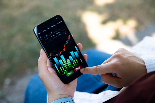 Woman is sitting on a bench, holding a smartphone displaying stock market data, rising chart and pressing the sell button. Online trading, investment, stock prices, financial advisor, assets, wealth.
