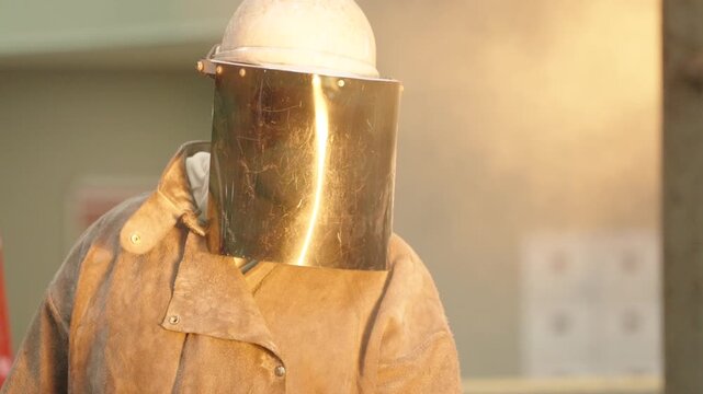 Operator in a heat resistant coat and reflective visor stands still inside a bright engineering steel foundry.