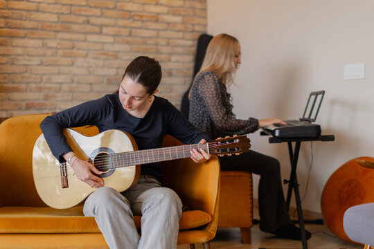 Teen girls making music indoors at home, one focused on guitar and the other on keyboard. Cozy living room with brick wall and warm furniture. Creativity, bonding, teamwork and leisure concepts. 