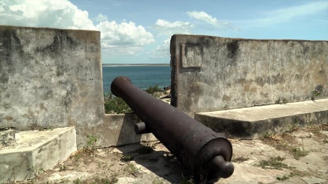 A weathered historical cannon stands guard on the weathered stone ramparts of Fort S&atilde;o Sebasti&atilde;o on Mozambique Island, overlooking the turquoise Indian Ocean under a cloudy sky.