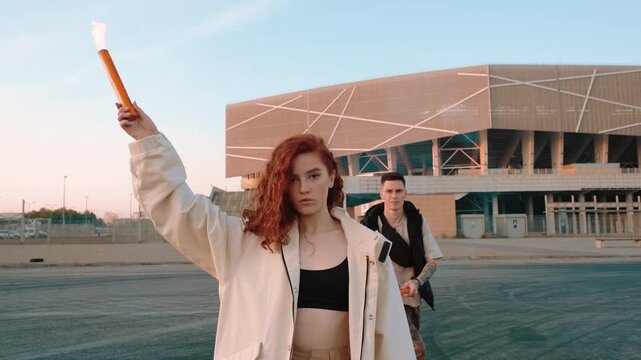 Red haired woman stands foreground with arm lifted, holding bright flare before large arena at dusk, while man behind mirrors defiant stance, with woman, flare, stadium, man detail