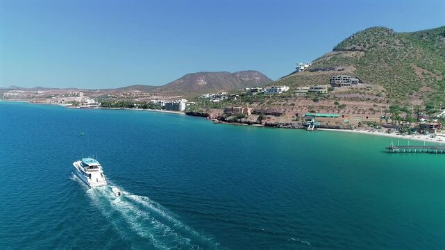4K aerial drone view follows a yacht over the water past Playa El Coromuel, with green desert hills and scattered houses along the coastline.