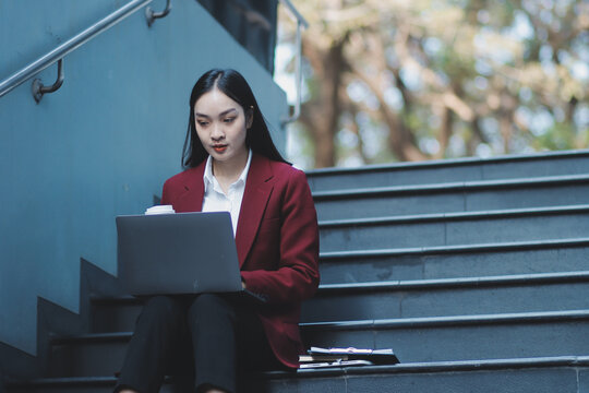 Focused woman in blazer types on laptop outdoors. Urban professional works remotely on stairs. She uses modern tech for business and connection.