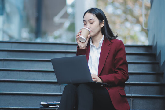 Focused woman in blazer types on laptop outdoors. Urban professional works remotely on stairs. She uses modern tech for business and connection.