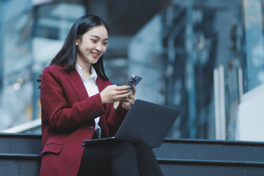 Focused woman in blazer types on laptop outdoors. Urban professional works remotely on stairs. She uses modern tech for business and connection.