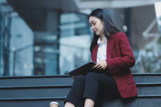 Focused woman in blazer types on laptop outdoors. Urban professional works remotely on stairs. She uses modern tech for business and connection.
