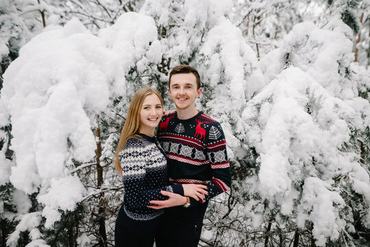 Happy couple in winter sweaters posing against backdrop of snow-covered spruce trees in fairytale forest. Romantic moment, valentines day.