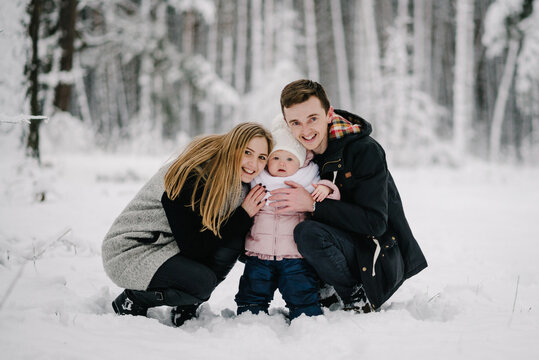 Happy parents hugging little daughter who stands in the center. The whole family smiling, looking at the camera, during winter walk in snowy forest. Warm emotions and family idyll. Close up.