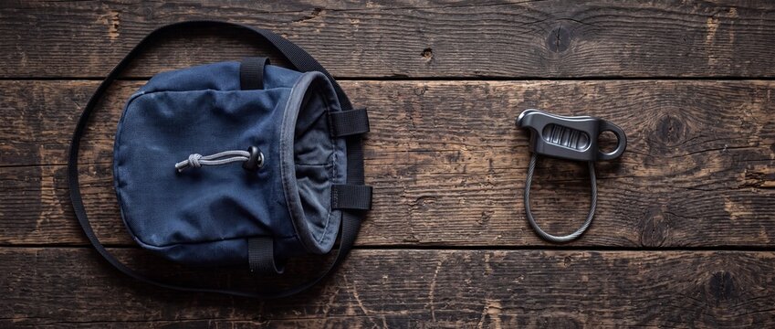 Navy blue chalk bag with drawstring and metal belay device arranged on dark rustic wooden background for climbing preparation