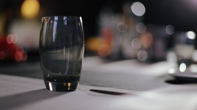 Gloved hand gently sets dark drinking glass on fabric-covered tabletop adjacent to rectangular slot, contact between glass base and woven fabric is visible as hand retracts, leaving glass standing