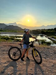 A boy wearing a helmet stands with his mountain bike on a rocky dirt path, looking out over a river valley toward distant mountains at sunset. The scene captures the spirit of active outdoor adventure © happy_artist