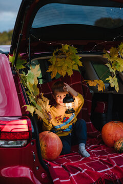 Kid having fun outdoors with pumpkin and plaid blanket. Smiling little boy holding lantern and leaves in car trunk decorated for autumn.