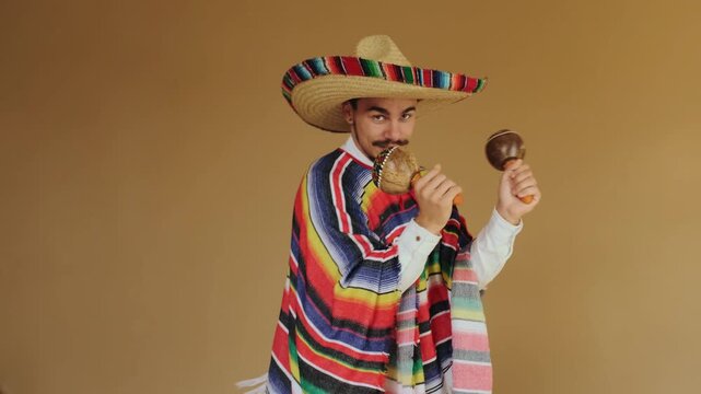 Midshot of young man wearing large woven straw sombrero and brightly striped wool serape shaking pair of wooden maracas with both hands, body rotated to right with shoulders and torso aligned to
