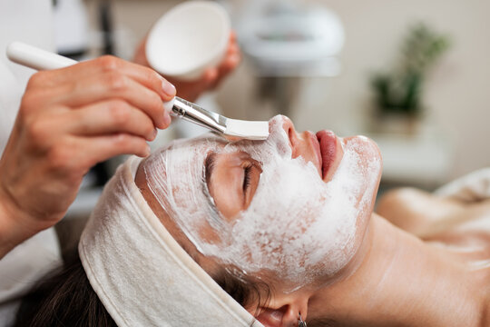 Professional aesthetician using a cosmetic brush to apply rejuvenating facial treatment in a modern wellness center.