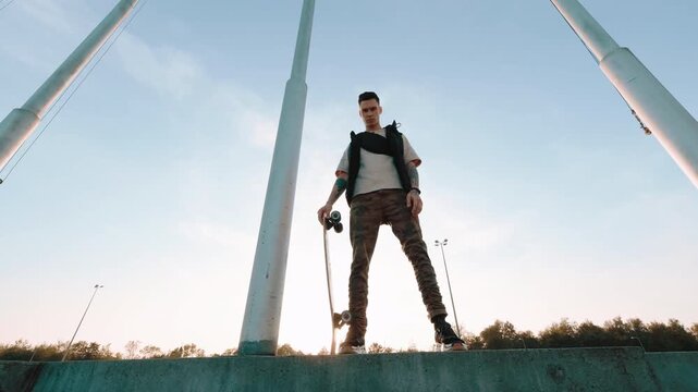 Young male skateboarder stands on concrete ledge with board beside body, framed by tall white poles against clear blue sky, with skateboarder, young, standing, male detail, showing outdoors, lowangle