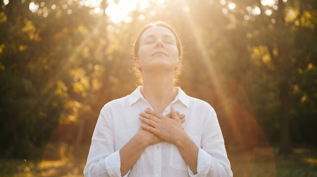 Grateful woman holding hands on chest with closed eyes under bright sunlight