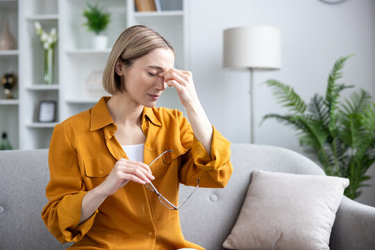 Woman sitting on a sofa at home, feeling a headache and eye strain, massaging her temples while holding glasses, reflecting discomfort and health issues