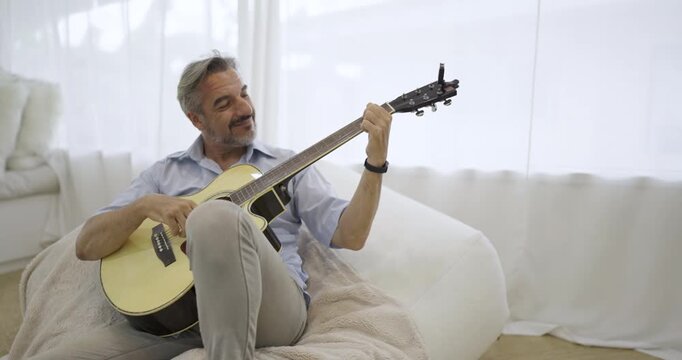 Portrait of elderly caucasian man musician as hobby. Senior male blogger teach guitar playing at living room. Handsome mature old man in casual clothes is smiling while playing guitar at home.