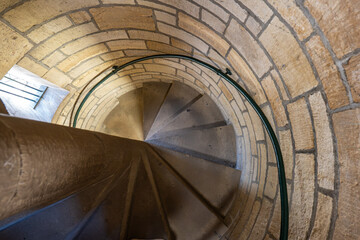 Naklejka premium Looking down a winding stone spiral staircase inside a historic tower with a metal handrail and stone wall.