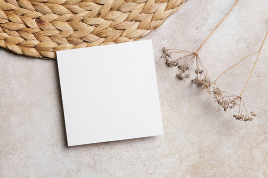 Blank white square card mockup placed on a textured beige surface with a woven straw mat and dried flowers, copy space for card design
