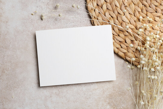 Horizontal blank white card on textured stone surface with delicate dried flowers and a natural woven placemat. Minimalist rustic flat lay design