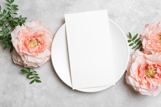 Blank vertical card mockup on a white plate with peach roses and green leaves on a gray concrete background. Elegant flat lay for menu, invitation, or greeting card designs