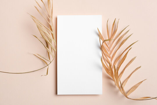 Vertical white blank card placed between two dried palm leaves on a beige background, ideal for elegant wedding invitations, menus, or minimalist designs