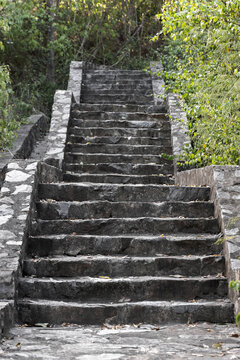 Weathered stone steps climb through dense green foliage, forming an outdoor staircase path that evokes exploration, solitude and history. Rough texture, fallen leaves and rustic walls