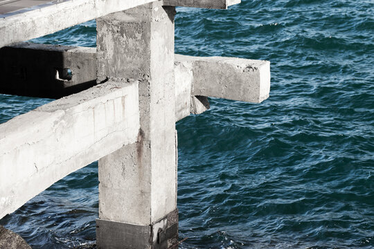 A weathered concrete pier and support columns extend over deep blue ocean waves, highlighting rugged texture, coastal structure and a moody maritime atmosphere suitable for seascape architecture