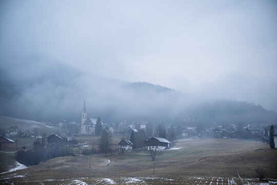 Wide shot of foggy alpine village with tall church spire surrounded by mist covered hills and light snow in winter. Atmospheric winter landscape in Gosau, Austria.