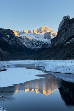 Cinematic sunset over Alpine lake in winter mountains. Winter mountain landscape. Golden hour reflection in calm mountain lake. Alpine peaks illuminated by orange light of setting sun.