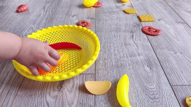 Child collects colorful toy food into a bright yellow bowl on a gray wooden floor, educational pretend play and early childhood development.