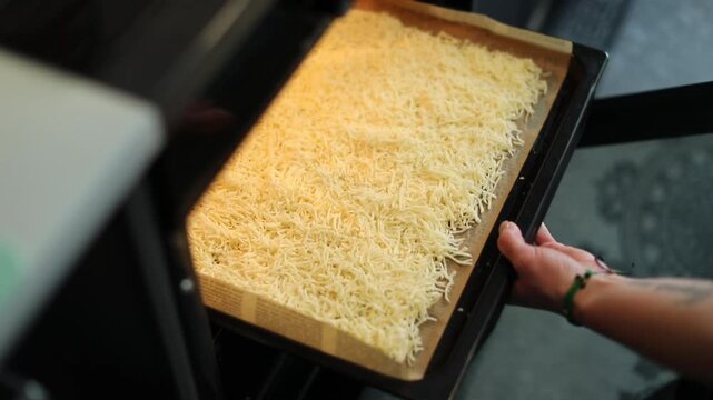 Woman placing a baking tray with grated cheese into a hot oven in a home kitchen. 
