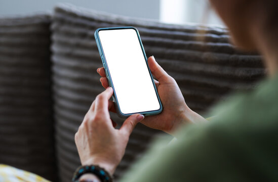 Woman Sits On The Couch At Home, Holding A Smartphone With A Blank White Screen In Hands