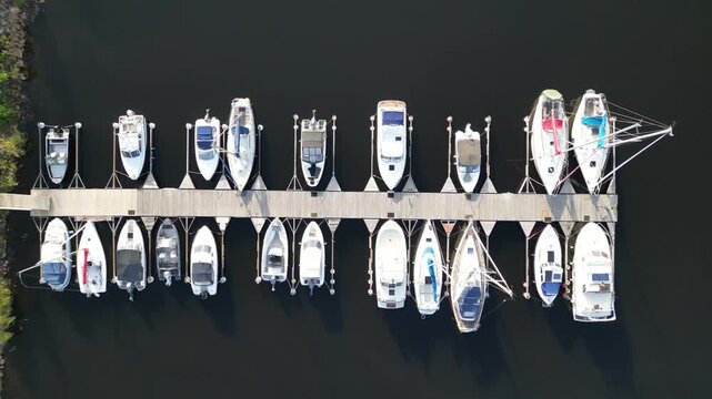 A striking top-down, bird's-eye perspective drone video showcasing motorboats and sailboats neatly docked along a wooden pier in a marina harbor.