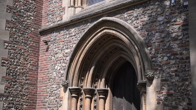 Sunlit Gothic side entrance of St Albans Cathedral reveals detailed stone arches and flint brickwork in a slow upward move.