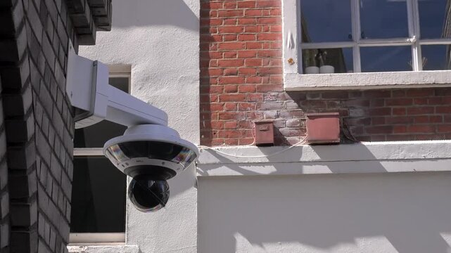 Static dome security camera hangs from a brick and plaster office wall in bright daylight, emphasizing surveillance and monitoring.
