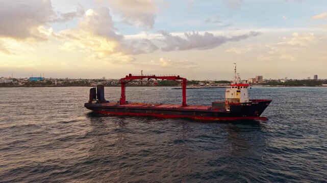 High-angle aerial drone orbiting right around an anchored cargo ship in the Caribbean Sea near the Santo Domingo coastline at dramatic sunset